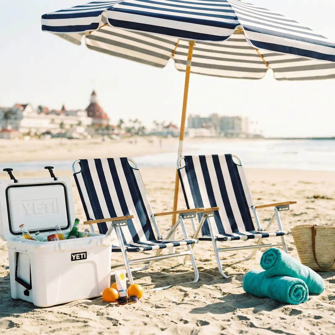 Beach day setup on Coronado sand with two striped lounge chairs, a white cooler, rolled teal towels, and a beach umbrella, with the Hotel del Coronado in the background.