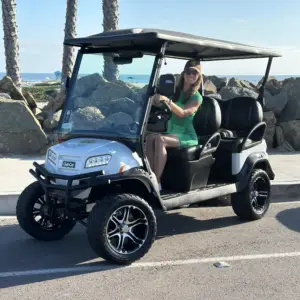 A woman in a green dress and baseball cap smiling from the driver's seat of a Nado Karts golf cart near the beach rocks.