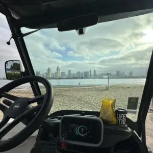 Driver's perspective from inside a luxury street-legal golf cart rental parked on a Coronado beach, looking across the bay at the downtown San Diego skyline with coffee on the dashboard.