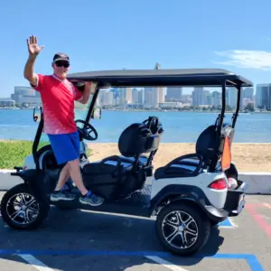 A man in a red shirt waving happily while standing on the side step of a Nado Karts golf cart near the bay.