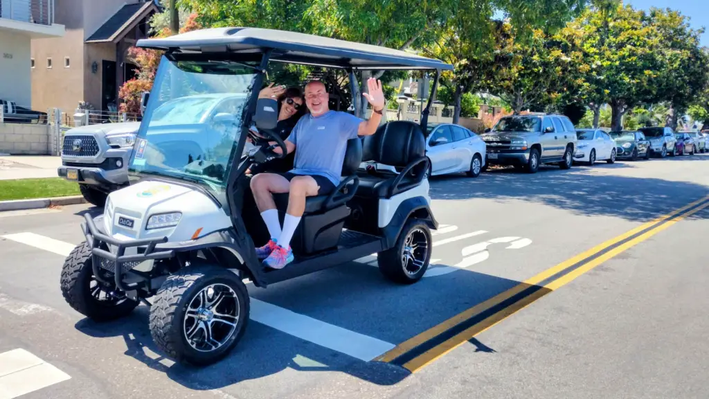 Two friends smiling and waving from a street-legal electric golf cart on a residential street in Coronado.