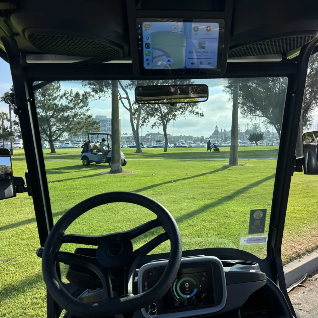 View from the driver's seat of a Nado Karts electric golf cart, showing the steering wheel and dual digital dashboard screens with Apple CarPlay, looking out toward the Hotel Del Coronado.
