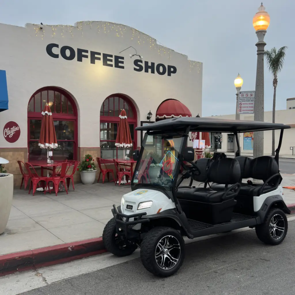 A white Club Car luxury street-legal golf cart rental from Nado Karts parked on Orange Avenue in front of a vintage coffee shop in Coronado, California.